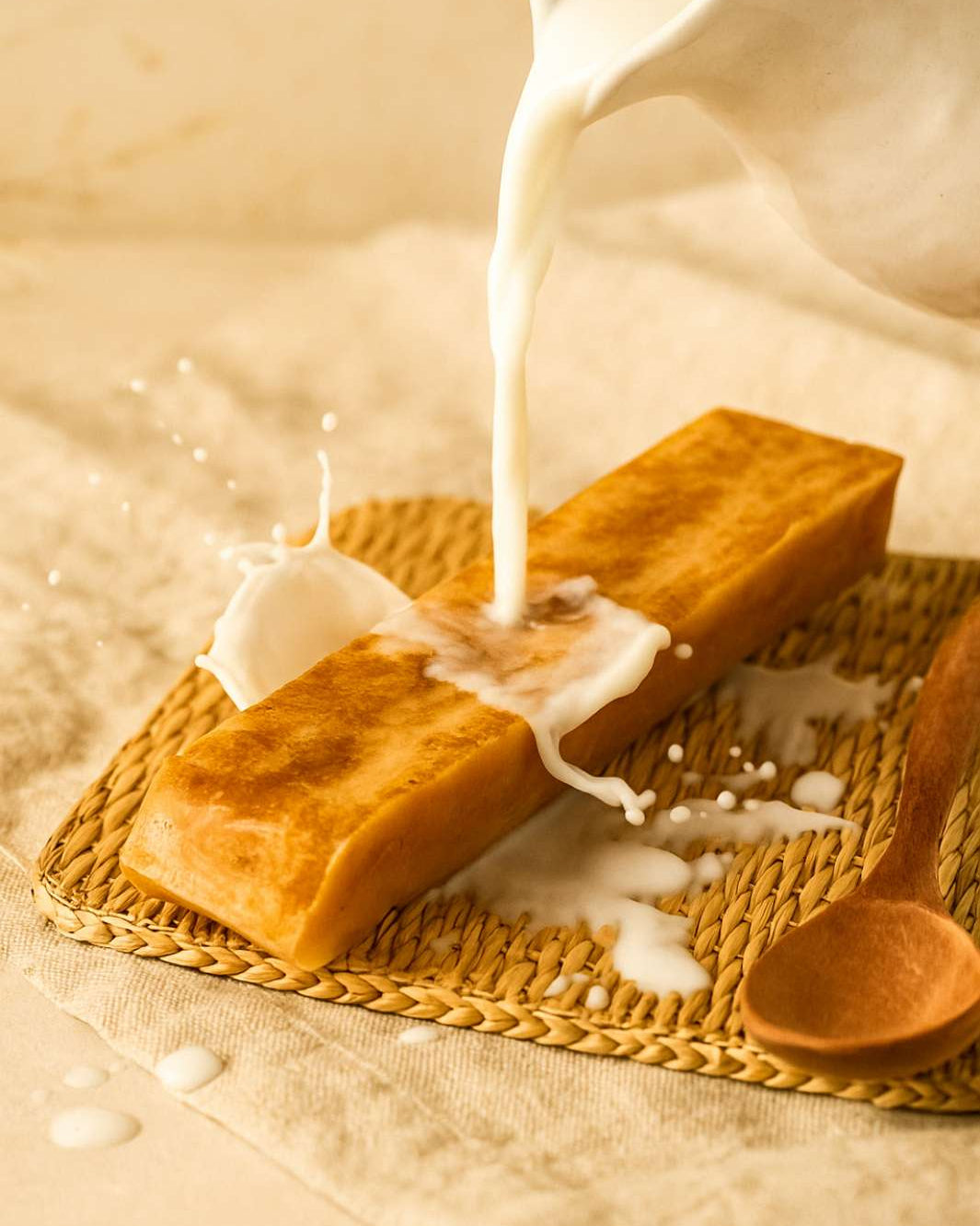 Two pieces of bread being drizzled with milk on a woven mat with a wooden spoon.